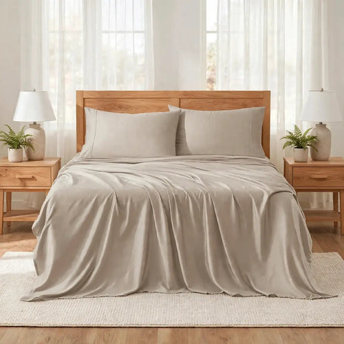 A neatly made bed with light beige or oatmeal-colored bedding, including a duvet cover and two matching pillowcases, styled in a bright bedroom with a wooden headboard, bedside tables, lamps, and plants.