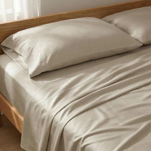 Beige bed sheets and matching pillowcases on a wooden bed frame, shown in a softly lit bedroom setting.