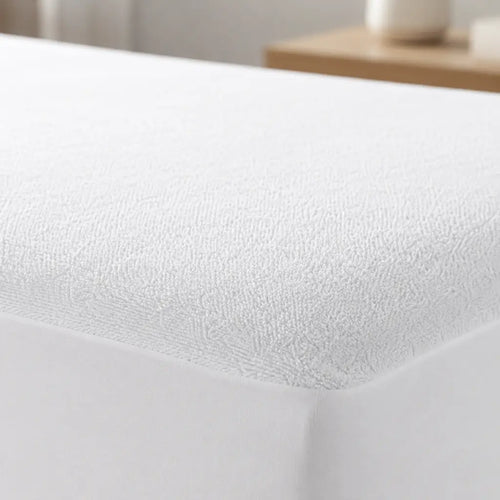 A close-up of a white fitted sheet with a textured, terry-like surface covering a mattress corner, with a softly blurred bedroom background.