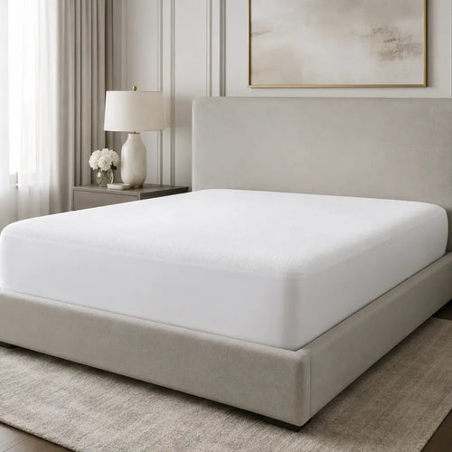 A white mattress fitted sheet shown on a bed frame in a softly styled bedroom with neutral walls, a bedside lamp, curtains, and framed wall art.