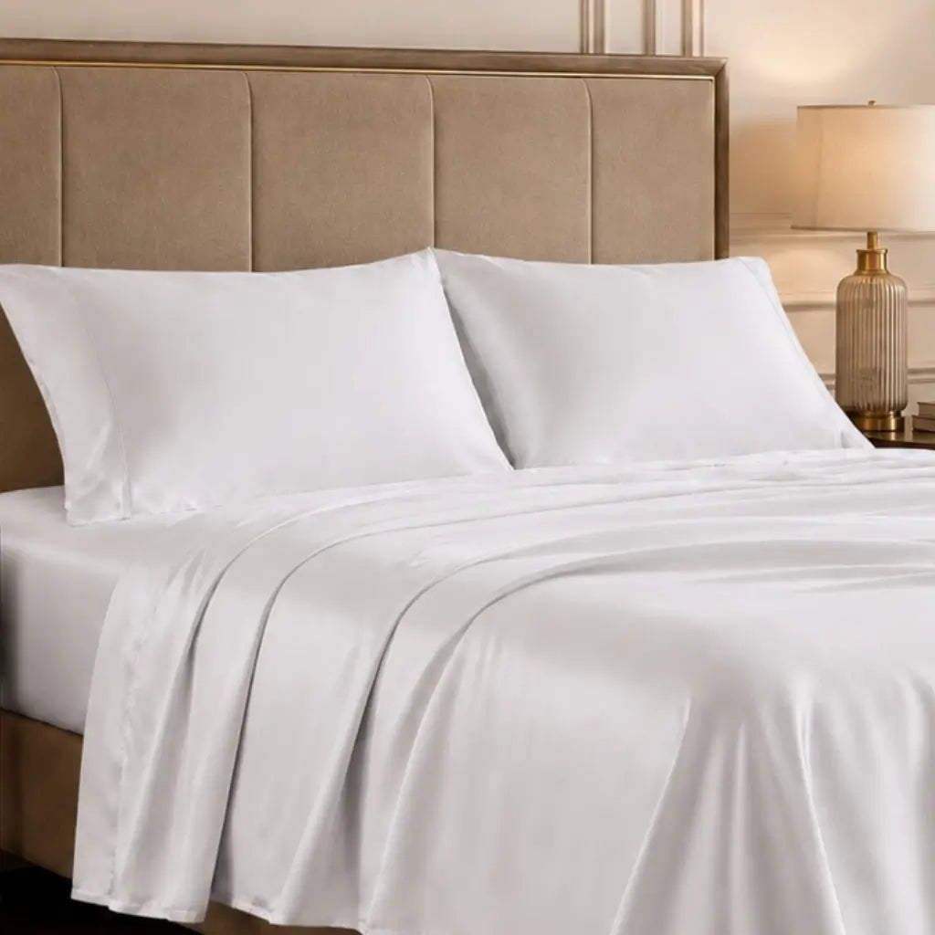 White satin sheet set with two matching pillowcases on a bed with a beige upholstered headboard and a bedside lamp.