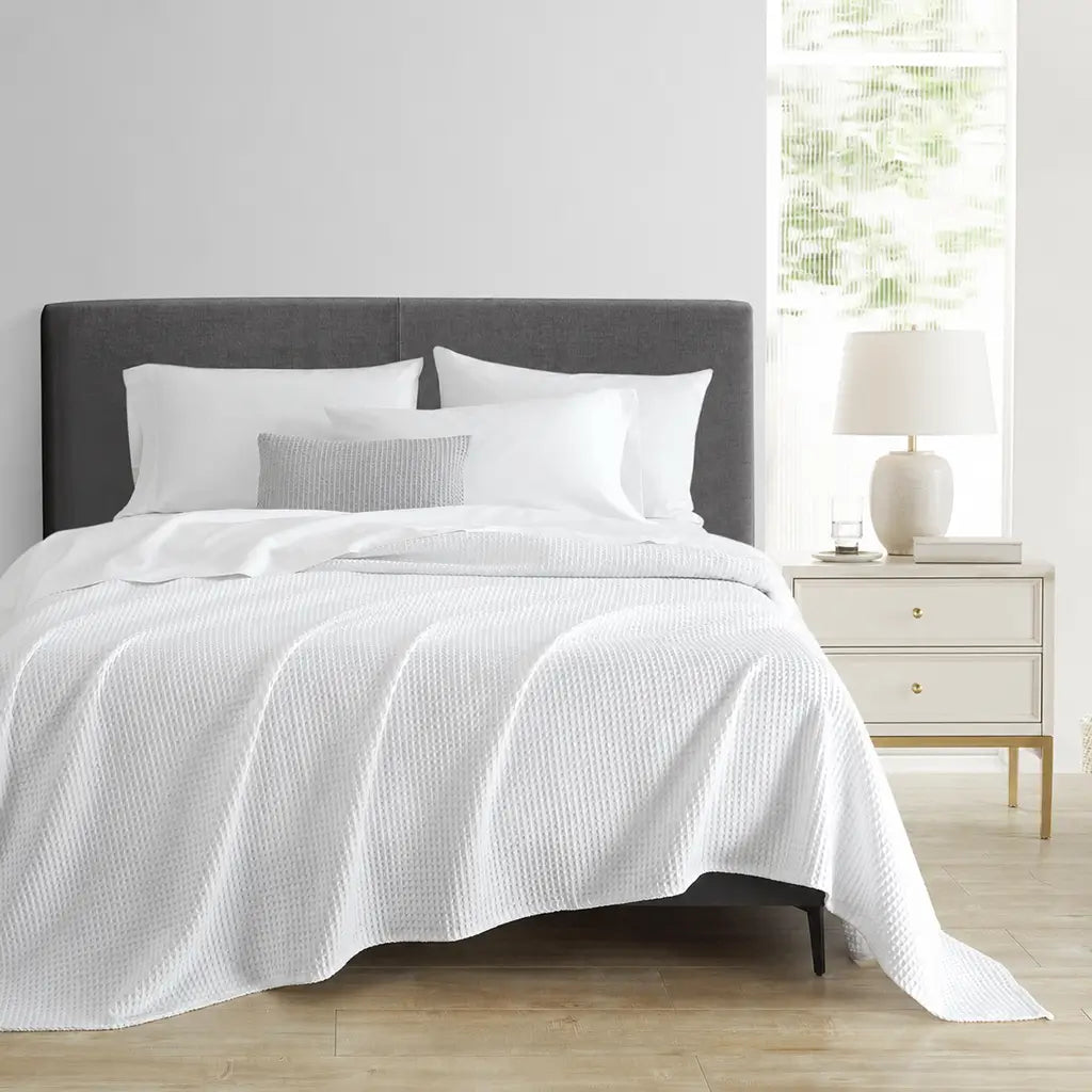 A neatly made bed with white textured bedding and pillows, set in a bright bedroom with a dark gray upholstered headboard, a white nightstand, and a lamp by a window.